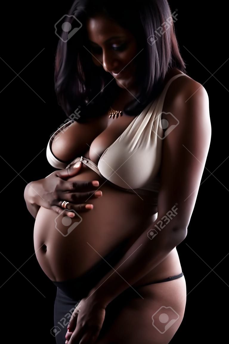 Young beautiful pregnant indian touching her tummy, over black background