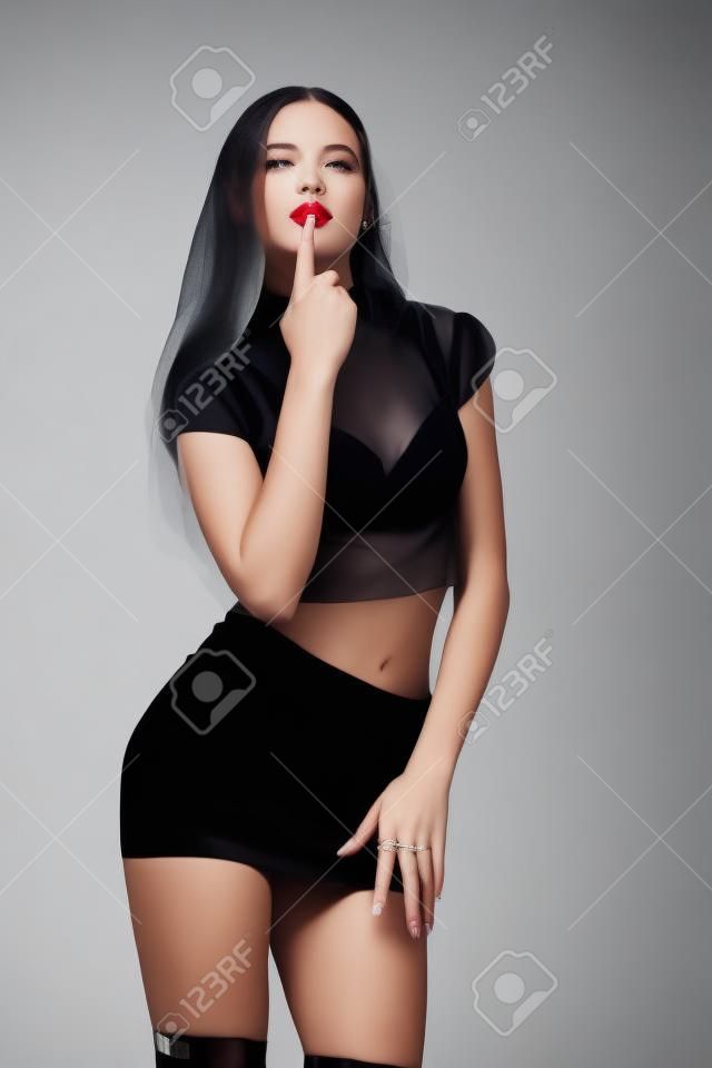 jeune femme de mode en talons hauts, studio tourné. Une femme vêtue d'une courte jupe noire et d'un haut transparent porta son doigt à ses lèvres.