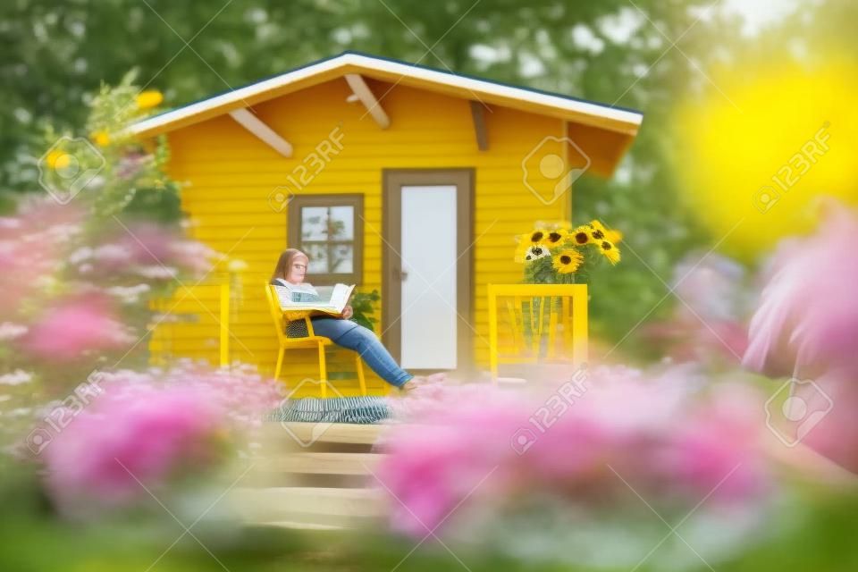 A teenage girl sitting in a chair on the veranda of a garden shed, reading a book. Lots of yellow coneflowers.