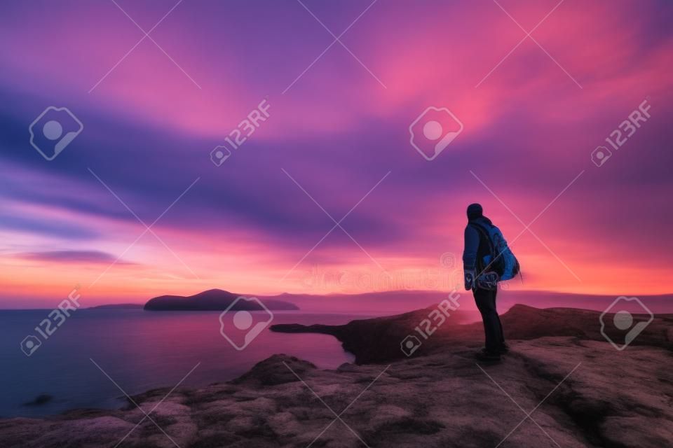 Young standing man with backpack. Hiker on the stone on the seashore at colorful sunset sky. Beautiful landscape with sporty man rocks sea and clouds at sunset. Sporty lifestyle
