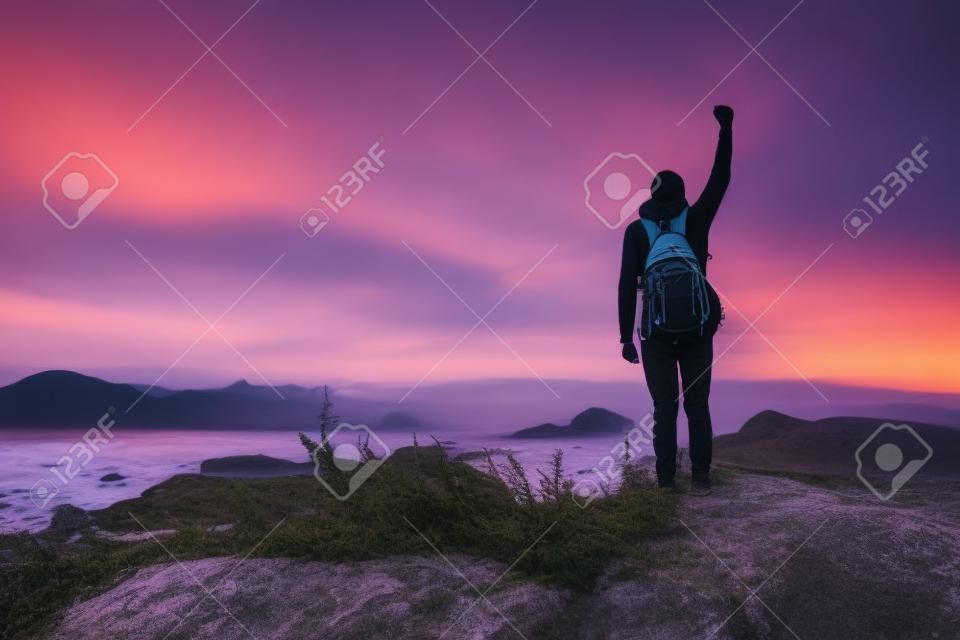 배낭을 메고 서 있는 젊은 남자. 다채로운 일몰 하늘에서 해변에 돌에 등산객. 스포티 한 남자 바위 바다와 일몰에 구름과 아름 다운 풍경. 스포티 라이프 스타일