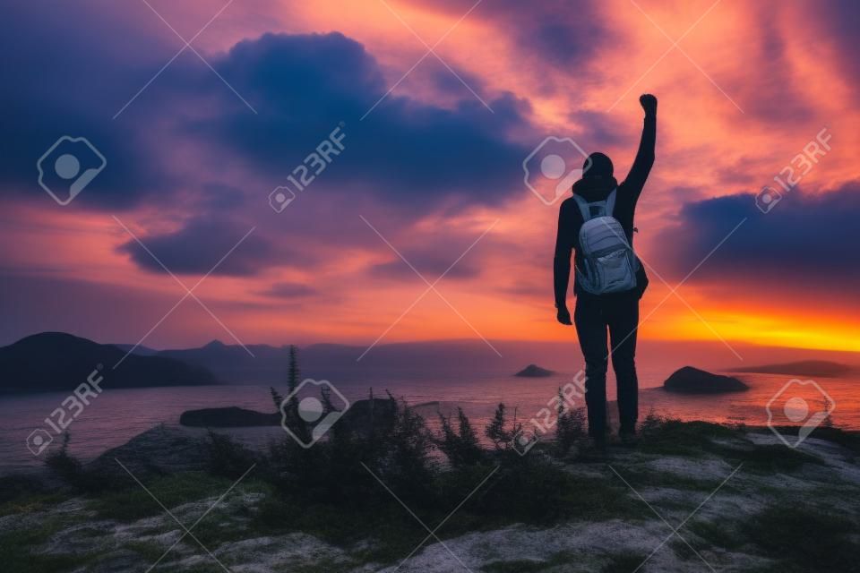 배낭을 메고 서 있는 젊은 남자. 다채로운 일몰 하늘에서 해변에 돌에 등산객. 스포티 한 남자 바위 바다와 일몰에 구름과 아름 다운 풍경. 스포티 라이프 스타일