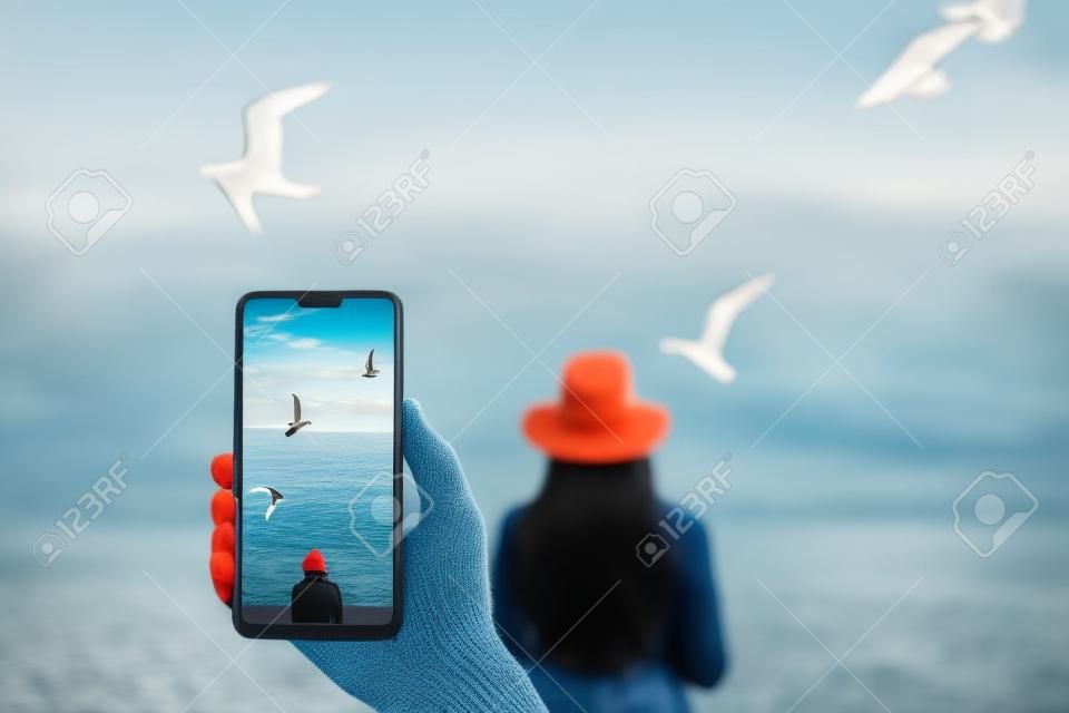 Um homem faz uma foto em um smartphone, menina olhando para o mar calmo no com o vento em seu cabelo e gaivotas no fundo.