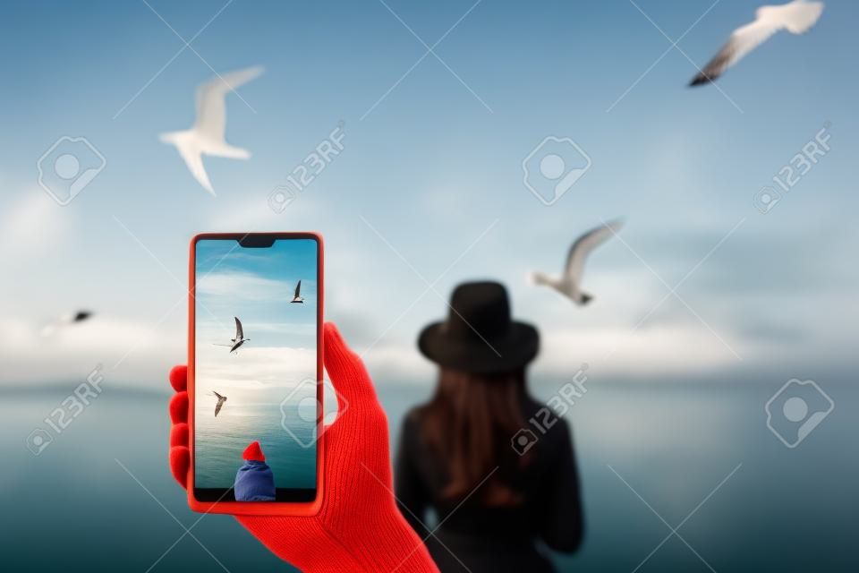 Un uomo fa una foto su uno smartphone, ragazza che guarda il mare calmo con il vento tra i capelli e i gabbiani sullo sfondo. Autunno nel cappello