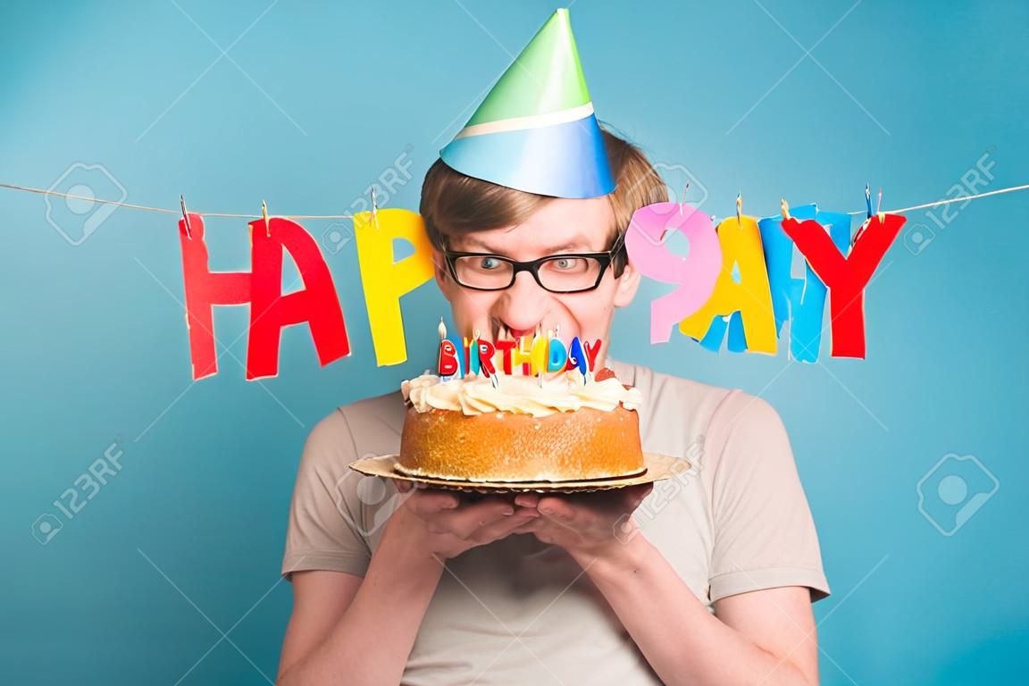 Il giovane pazzo divertente con un cappello di carta di auguri vuole mordere un pezzo di torta di congratulazioni. Concetto di scherzare e auguri di compleanno. Copia spazio