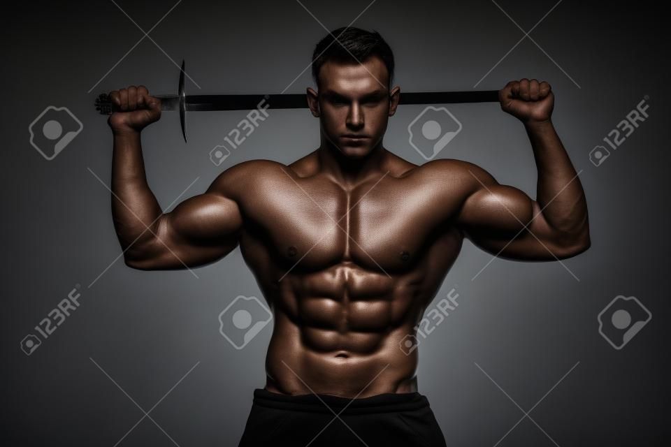 Homme bodybuilder posant avec une épée isolée sur fond noir. Homme torse nu sérieux démontrant son corps masculin