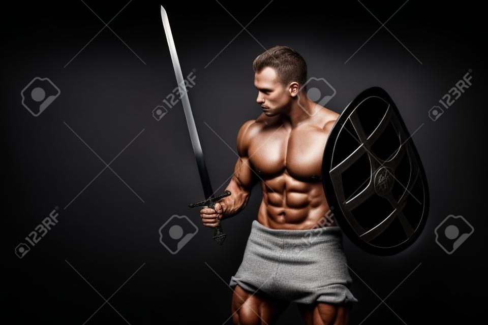 Bodybuilder man posing with a sword and shield isolated on black background. Serious shirtless man demonstrating his mascular body