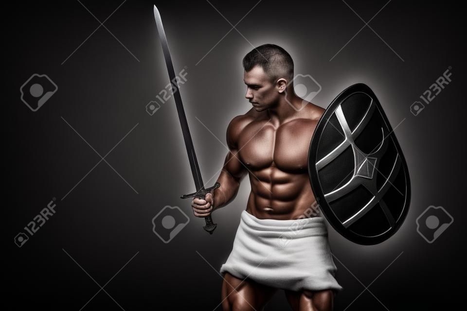 Bodybuilder man posing with a sword and shield isolated on black background. Serious shirtless man demonstrating his mascular body