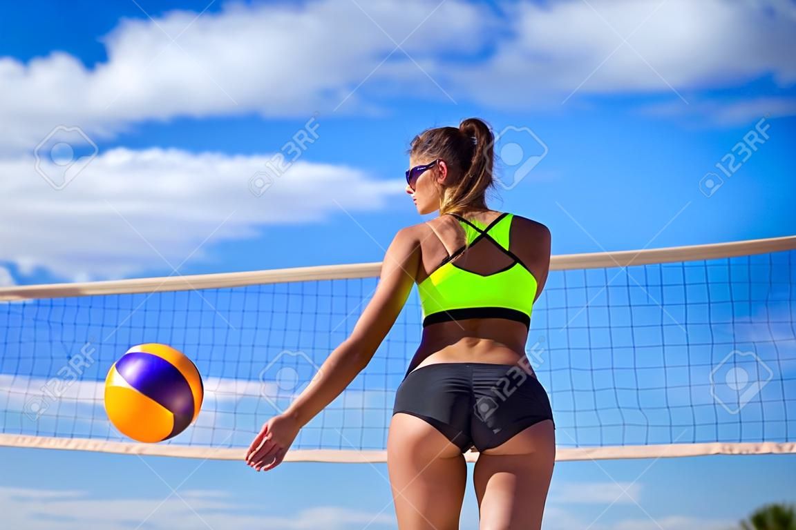 Vue arrière d'une jeune fille athlétique debout sur la plage sur fond de filet et de ciel avec un ballon et une bouteille d'eau dans les mains