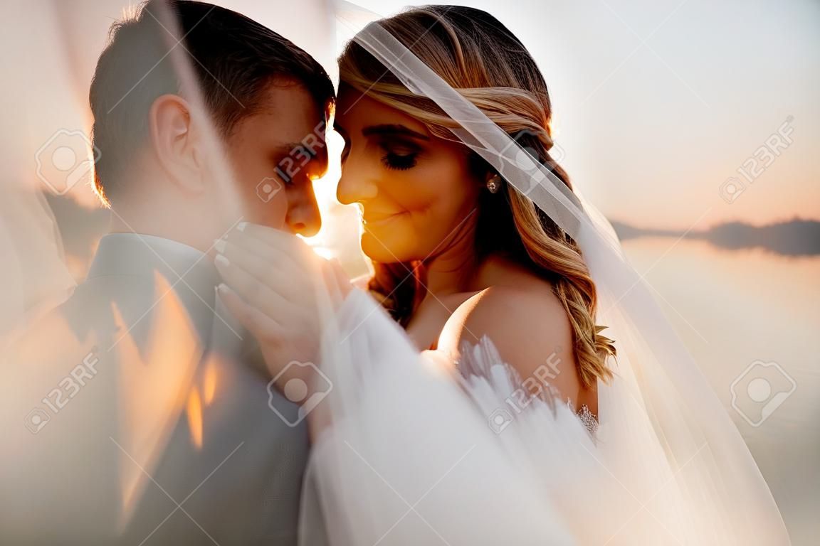 the bride and groom under the veil on the sunset by the river