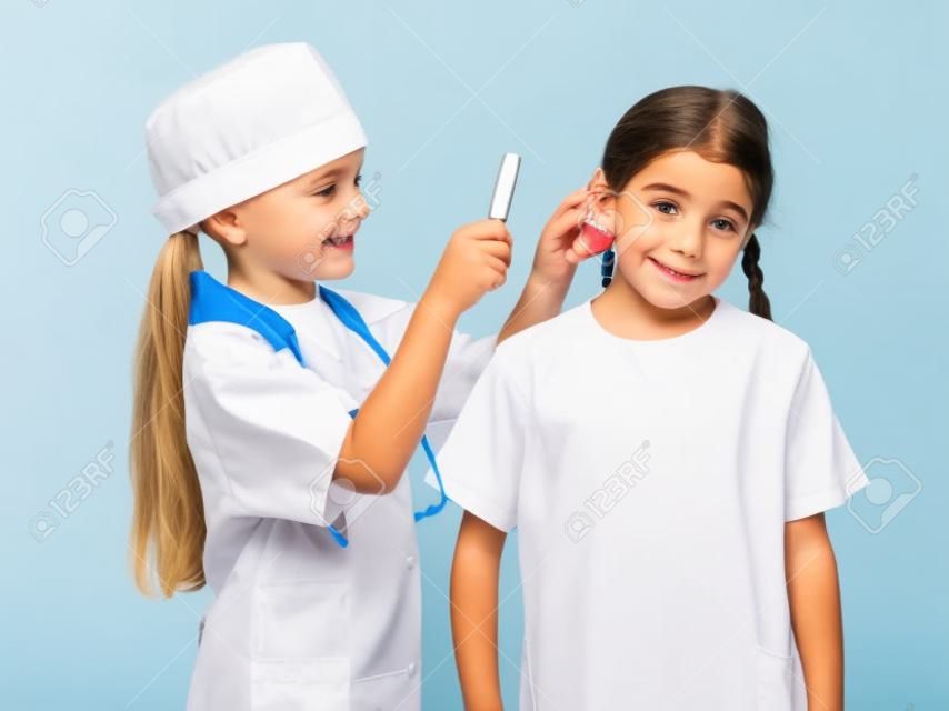 Little girl in doctor costume are inspecting the another girl s ears with magnifier, isolated on white