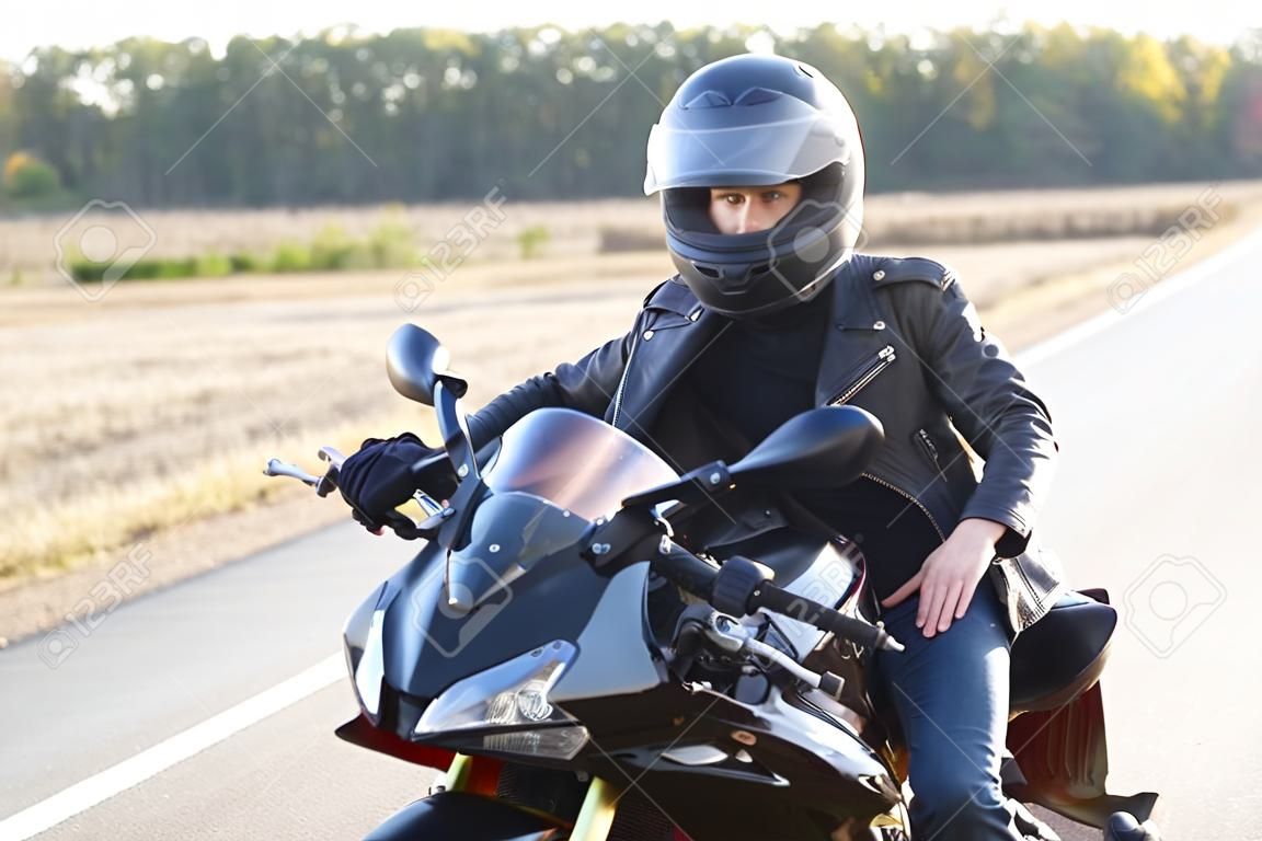 Street style concept. Active professional rider wears helmet, black leather jacket, poses on motorrbike at road, enjoys high speed, ready to cover long distance and have trip. Horizontal shot