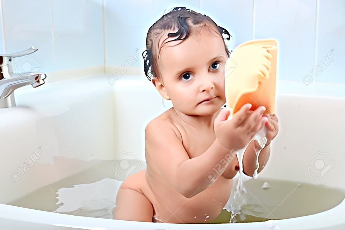 Beautiful infant sitting around foam in bathroom puts washcloth in two hands, trying to squeeze it, focused on washing process. Attractive little kid is curious about taking bath. Care concept.