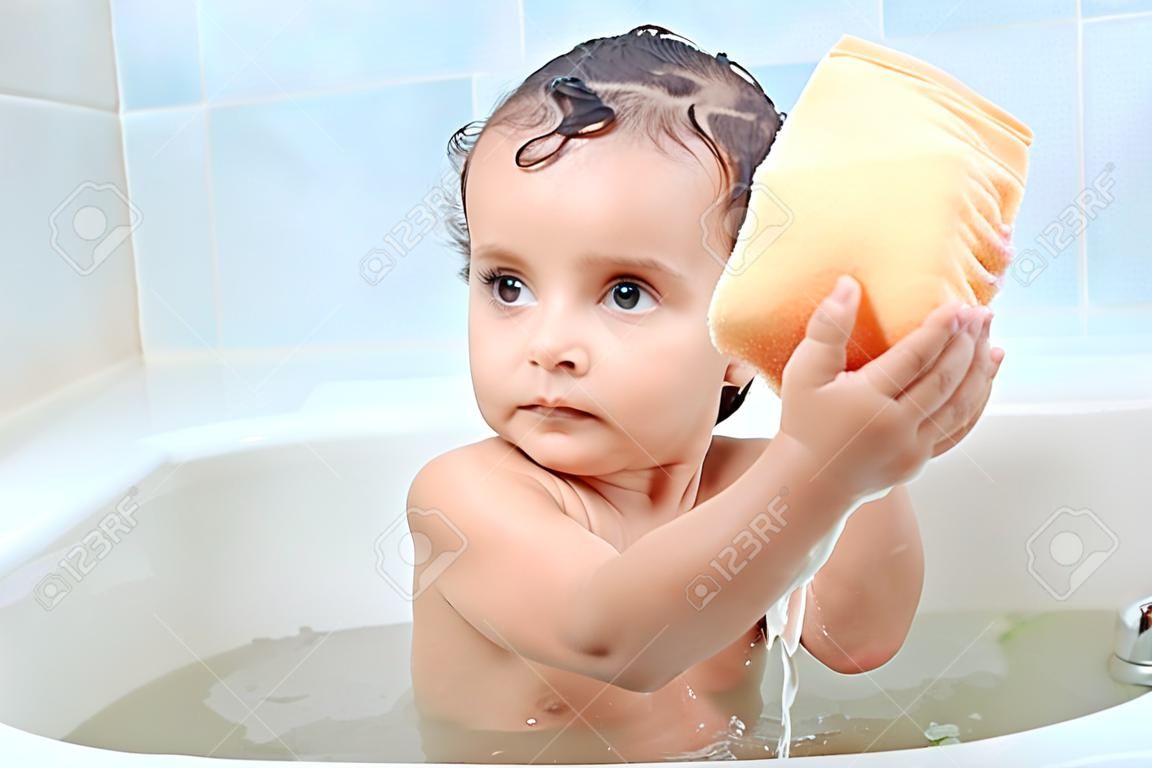 Beautiful infant sitting around foam in bathroom puts washcloth in two hands, trying to squeeze it, focused on washing process. Attractive little kid is curious about taking bath. Care concept.