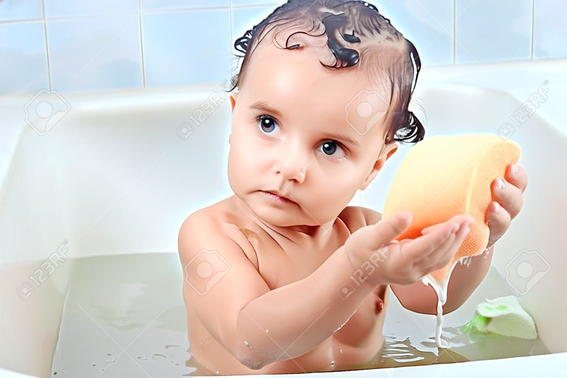 욕실에 있는 거품 주위에 앉아 있는 아름다운 아기는 수건을 두 손에 쥐고 짜려고 하고 세탁 과정에 집중합니다. 매력적인 어린 아이가 목욕에 대해 궁금해합니다. 케어 개념입니다.