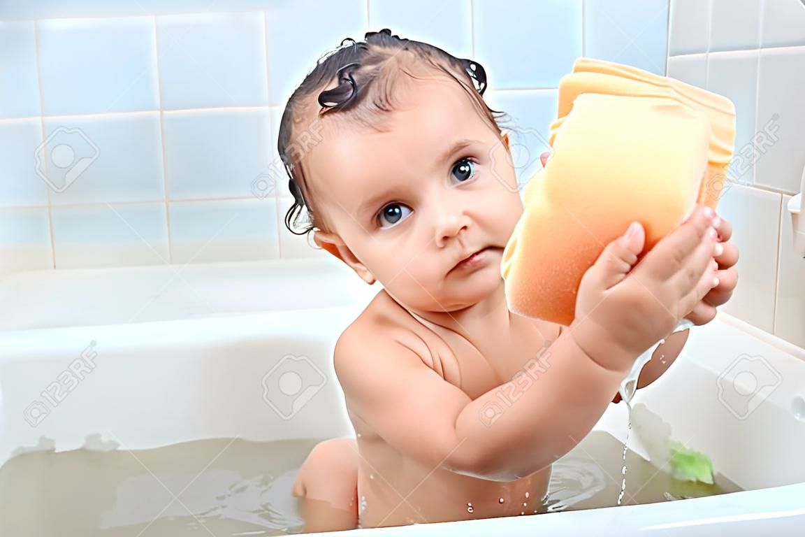 욕실에 있는 거품 주위에 앉아 있는 아름다운 아기는 수건을 두 손에 쥐고 짜려고 하고 세탁 과정에 집중합니다. 매력적인 어린 아이가 목욕에 대해 궁금해합니다. 케어 개념입니다.