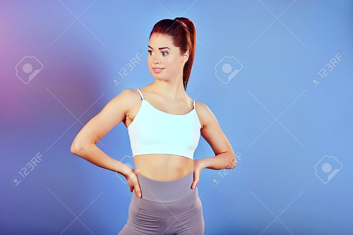 Portrait d'une jeune sportive caucasienne sportive et mince portant un soutien-gorge de sport blanc, debout avec les mains sur les hanches, regardant de côté, a une queue de cheval, copiez l'espace pour la publicité.