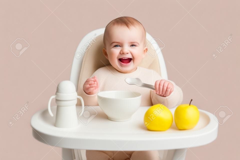 Imagem de deligted rindo bebê menina vestidos em jumper bege sentado em cadeira alta e comer, isolado sobre fundo branco, segurando colher nas mãos, desfrutando de purê de frutas saborosas.