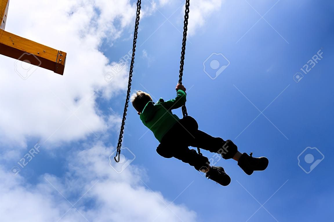 Silhouette of a boy swinging on a swing  against a deep blue sky with puffy clouds