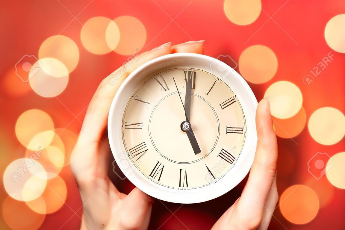 Female hands with cup and clock on color background with blurred lights