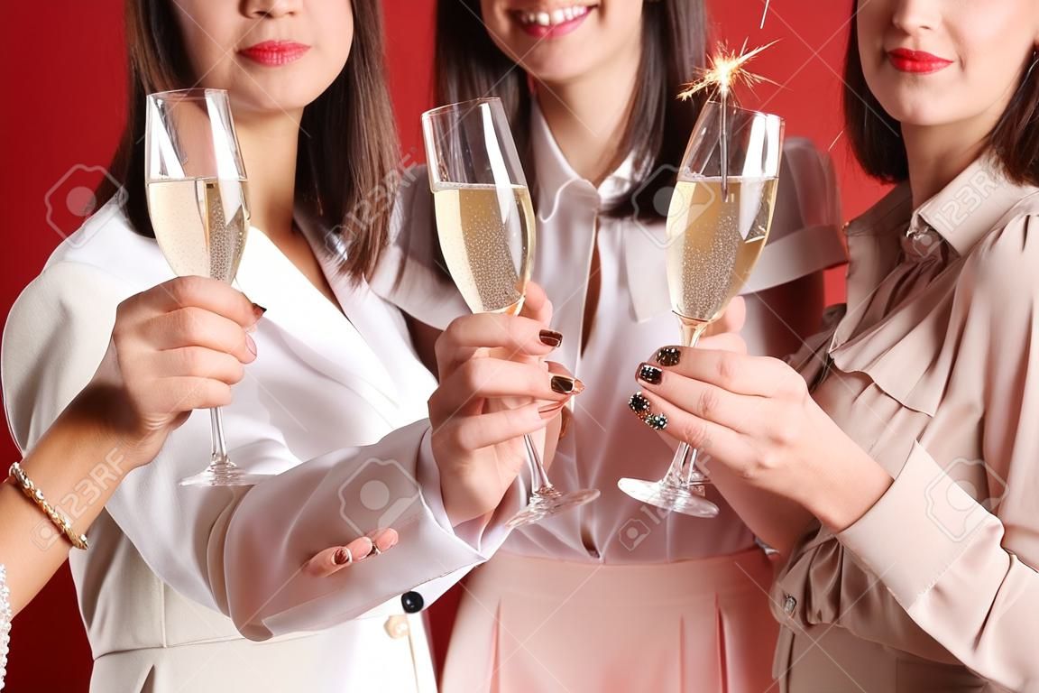 Beautiful young women with champagne and sparklers on red background, closeup. New Year celebration
