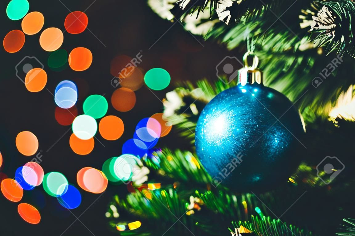 Blauer Weihnachtsball auf dem gezierten Weihnachtsbaum mit mehrfarbigem bokeh rundem Girlandenlichthintergrund