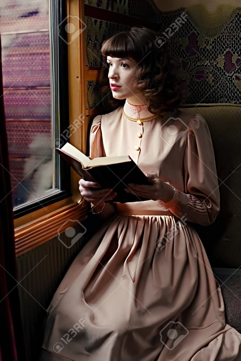 Mujer joven en vestido beige de época de principios del libro de lectura del siglo 20 en el coupé de tren de ferrocarril retro