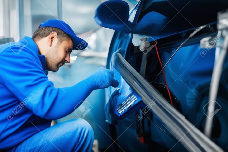 Widok z boku mechanika samochodowego w niebieskim mundurze i czapce naprawy przewodów elektrycznych w drzwiach starego samochodu. Mężczyzna w średnim wieku wymieniający uszkodzone detale na nowe w serwisie samochodowym