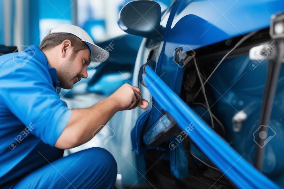 Zijaanzicht van auto monteur in blauw uniform en kap herstellen van elektrische draden in deur van oude auto. Middeleeuwse man die beschadigde details aan nieuwe in auto tankstation