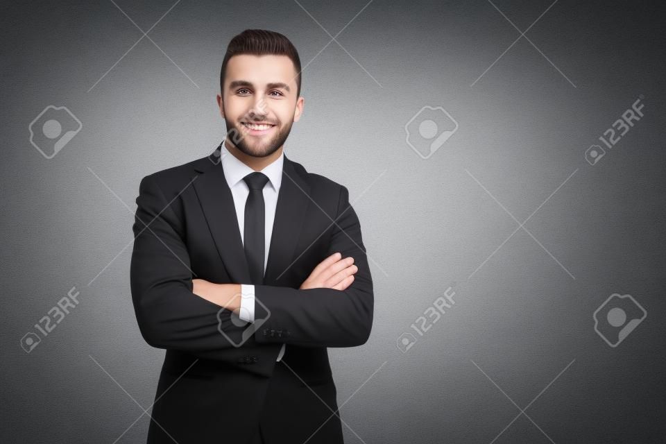 Vorderansicht eines jungen lächelnden Geschäftsmannes im schwarzen Anzug, der mit gekreuzten Händen posiert. Erfolgreiche brünette Arbeiterin steht und blickt in die Kamera, isoliert auf grauem Hintergrund. Geschäftskonzept.