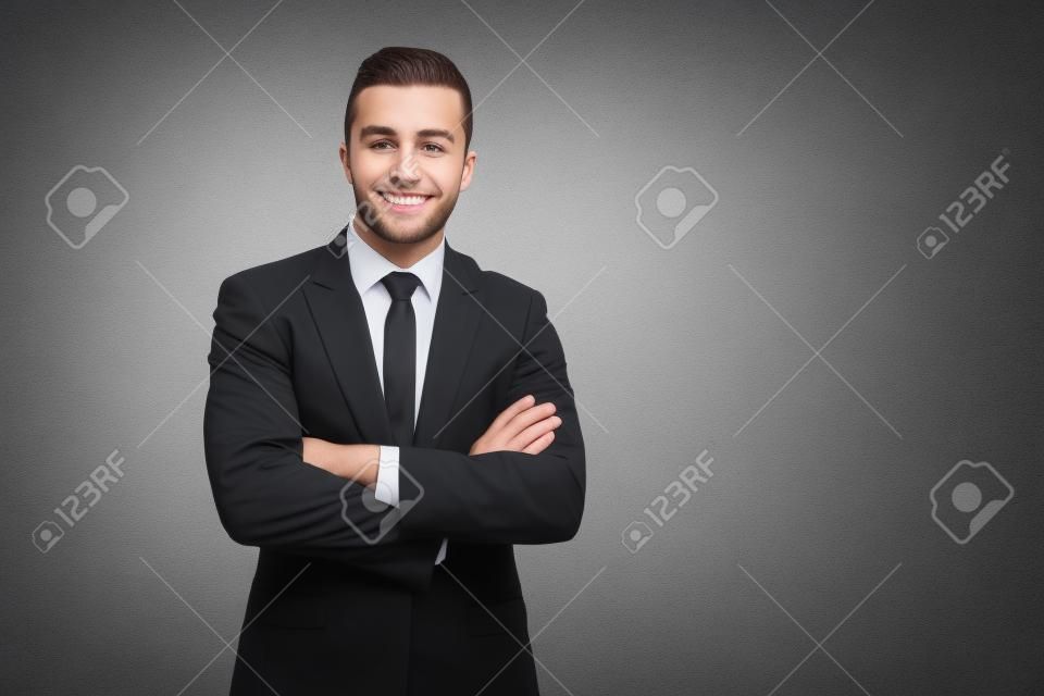 Vorderansicht eines jungen lächelnden Geschäftsmannes im schwarzen Anzug, der mit gekreuzten Händen posiert. Erfolgreiche brünette Arbeiterin steht und blickt in die Kamera, isoliert auf grauem Hintergrund. Geschäftskonzept.