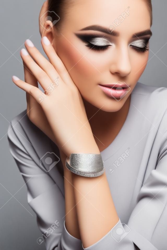 Crop de modelo feminino jovem com maquiagem perfeita apresentando pulseira de prata minimalista. Retrato de mulher posando em estúdio, isolado no fundo cinza. Conceito de joalheria.