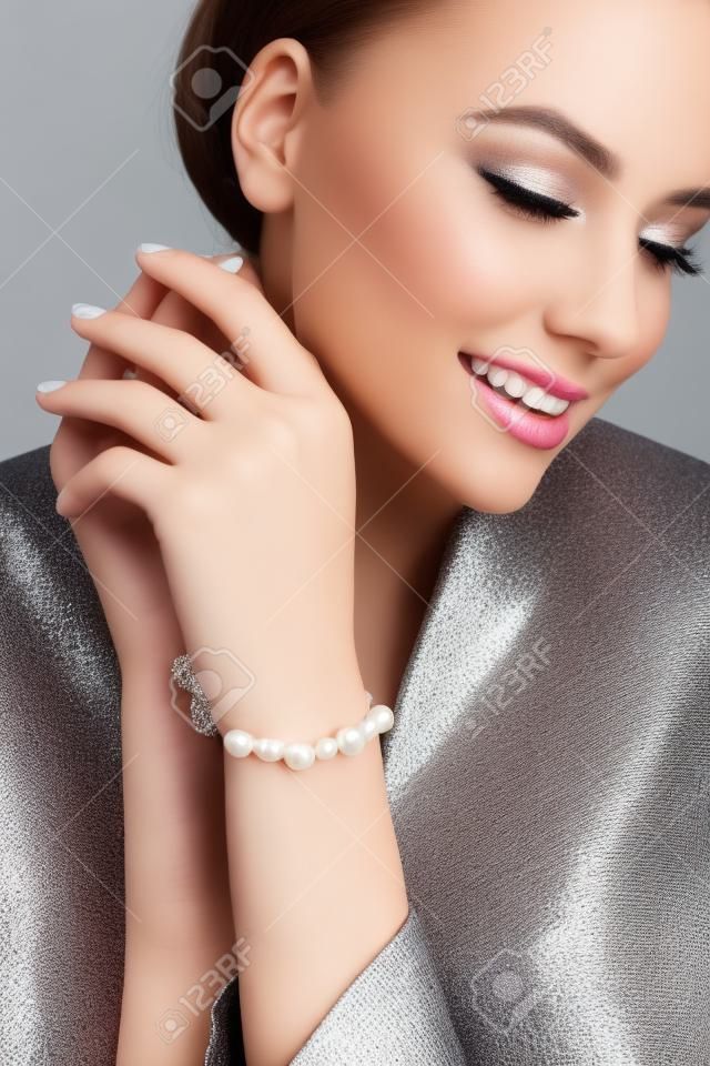 Bliska portret młodej pięknej uśmiechniętej modelki w formalnym garniturze prezentującej srebrną bransoletkę z białymi perłami. Kobieta z idealnym makijażem pozowanie w studio, na szarym tle.
