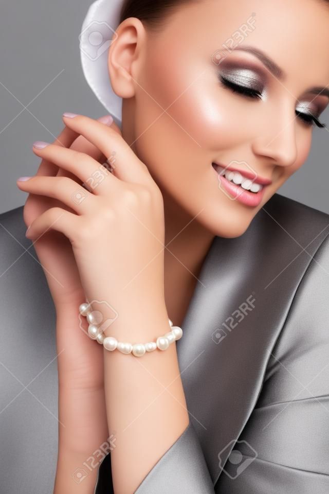 Close up portrait of young beautiful smiling female model in formal suit presenting silver bracelet with white pearls. Woman with perfect makeup posing in studio, isolated on gray.