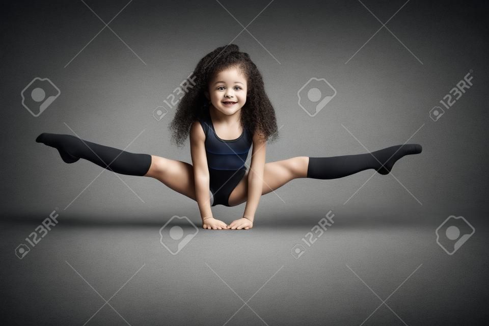 Kleine weibliche professionelle Turnerin, die Split macht, auf den Armen auf dem Boden stehend, isoliert auf grauem Studiohintergrund. Lächelndes Mädchen in schwarzer Sportbekleidung und Kniestrümpfen mit lockigem Haar, das Flexibilität zeigt.
