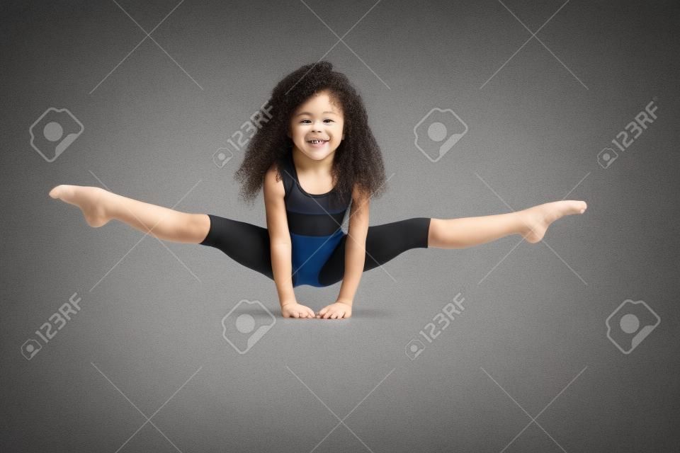 Pequeña gimnasta profesional femenina haciendo split, de pie sobre los brazos en el piso, aislado sobre fondo gris de estudio. Niña sonriente en ropa deportiva negra y calcetines hasta la rodilla con cabello rizado mostrando flexibilidad.