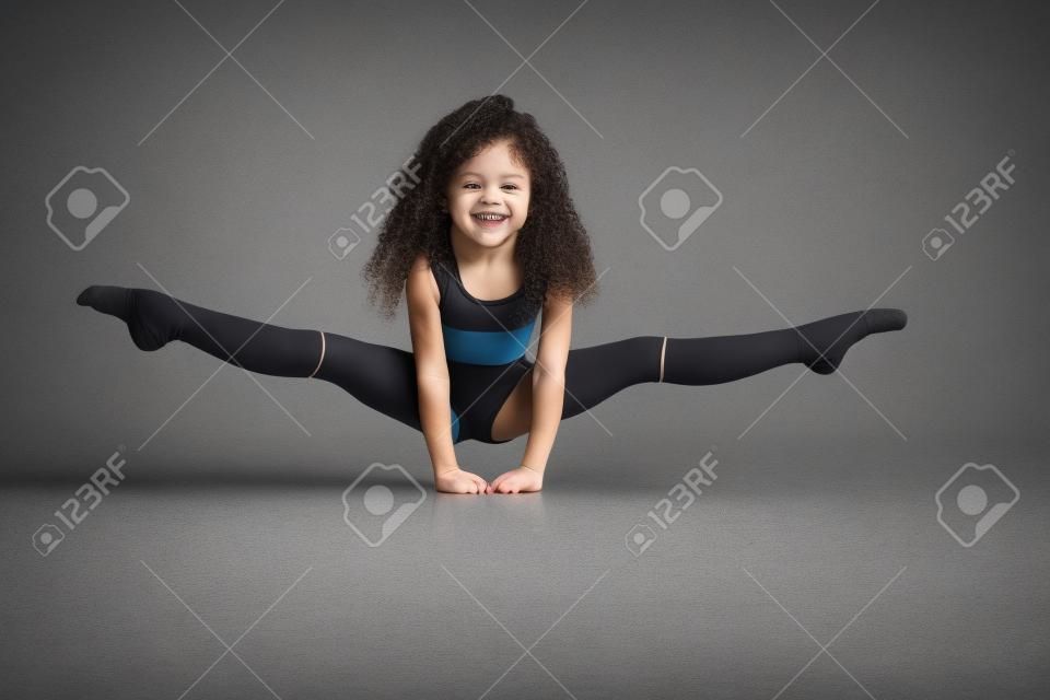 Kleine vrouwelijke professionele turnster doen split, staan op armen op de vloer, geïsoleerd op grijze studio achtergrond. Lachend meisje in zwarte sportkleding en knie sokken met krullend haar tonen flexibiliteit.