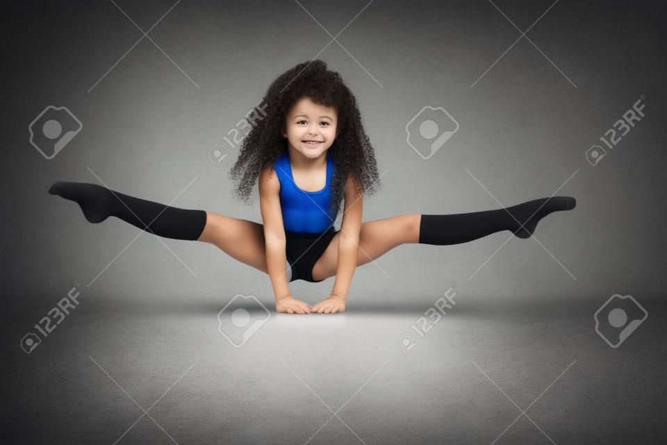 Kleine weibliche professionelle Turnerin, die Split macht, auf den Armen auf dem Boden stehend, isoliert auf grauem Studiohintergrund. Lächelndes Mädchen in schwarzer Sportbekleidung und Kniestrümpfen mit lockigem Haar, das Flexibilität zeigt.