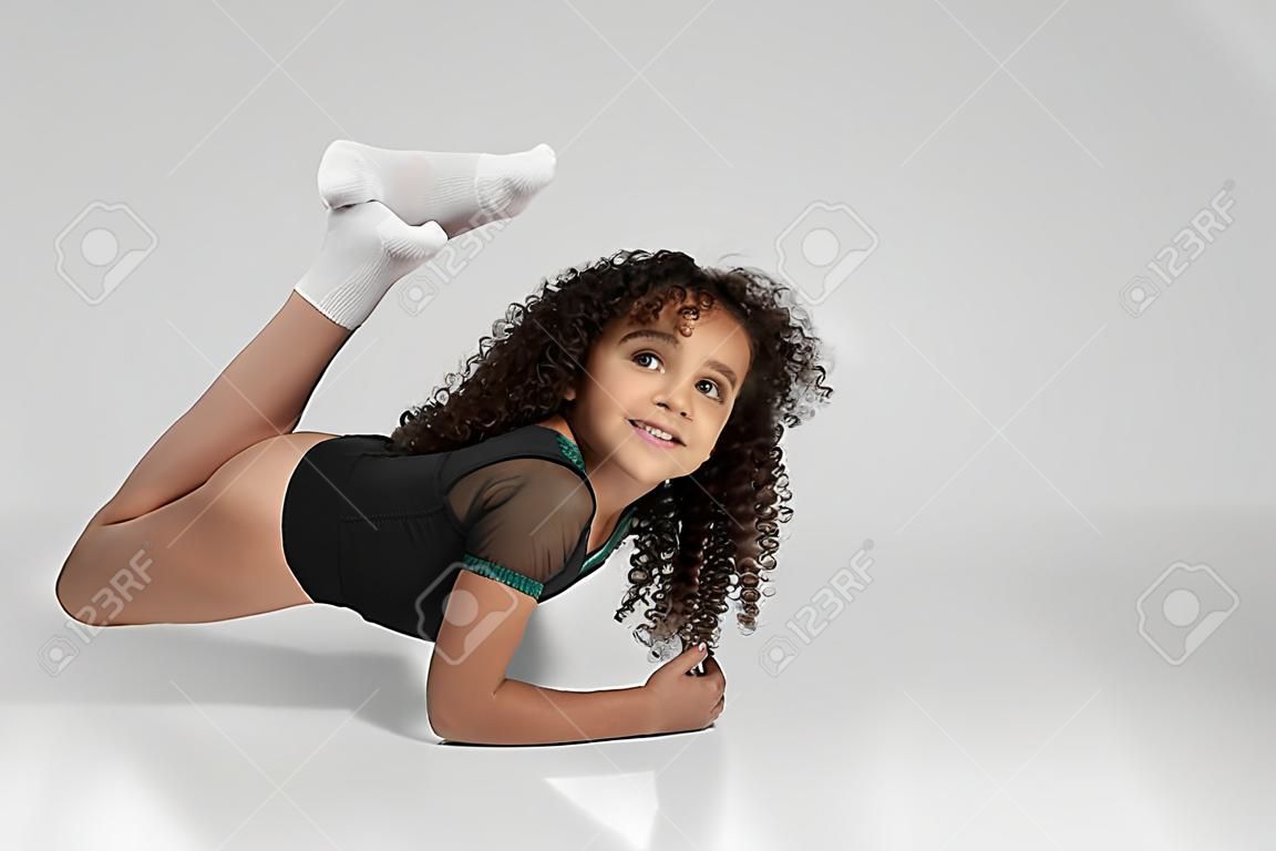 회색 배경에 격리된 보트 운동을 보여주는 운동복과 무릎 양말을 신고 웃는 귀여운 소녀. 카메라를 보고 유연성을 보여주는 곱슬 머리를 가진 작은 여성 전문 체조 선수.