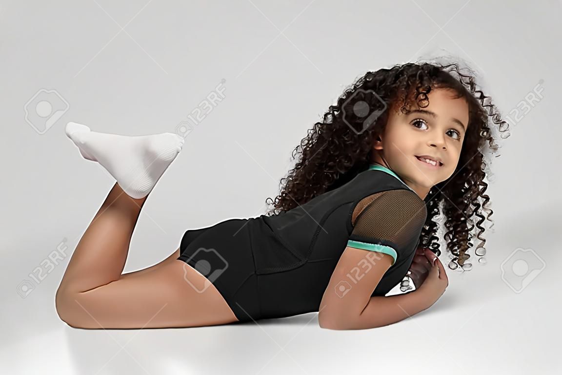 Nettes lächelndes Mädchen in Sportkleidung und Kniestrümpfen, die Bootsübungen demonstrieren, isoliert auf grauem Hintergrund. Kleine professionelle Turnerin mit lockigem Haar, die Flexibilität zeigt und in die Kamera schaut.