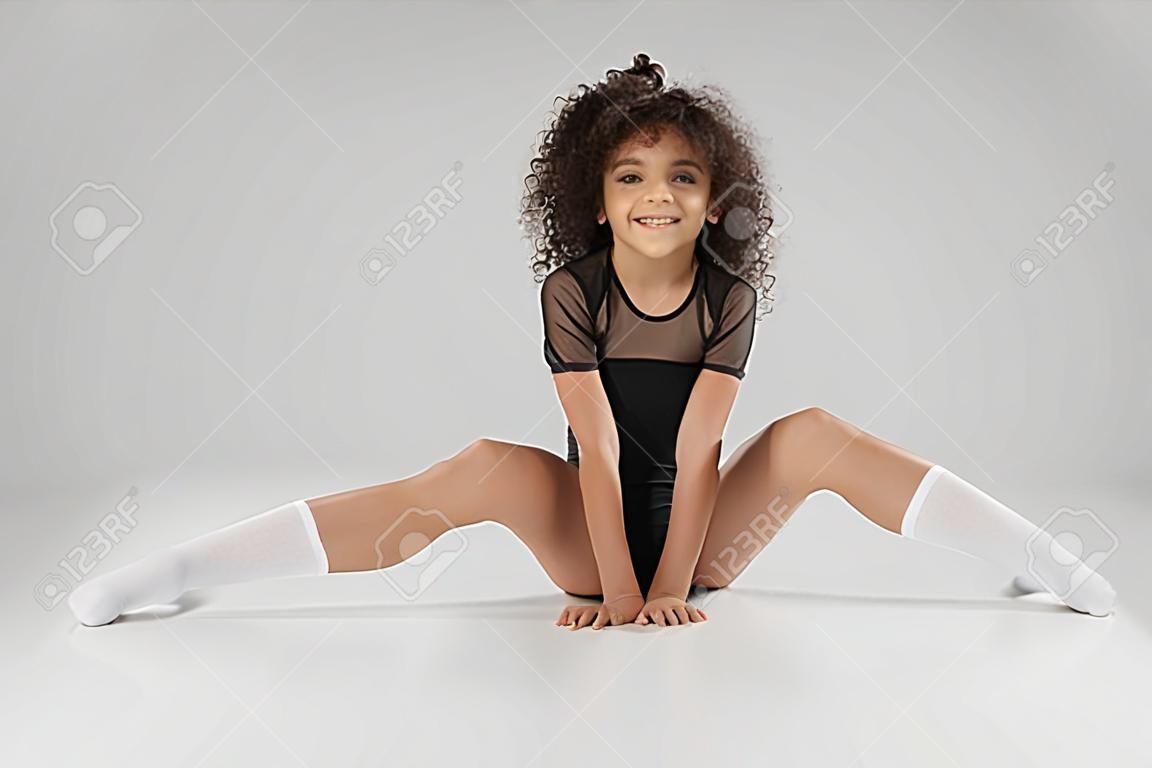 Lächelndes Mädchen in schwarzer Sportbekleidung und Kniestrümpfen mit lockigem Haar, das Flexibilität zeigt. Junge weibliche professionelle Turnerin sitzt auf dem Boden mit breiten Beinen, isoliert auf grauem Studiohintergrund.