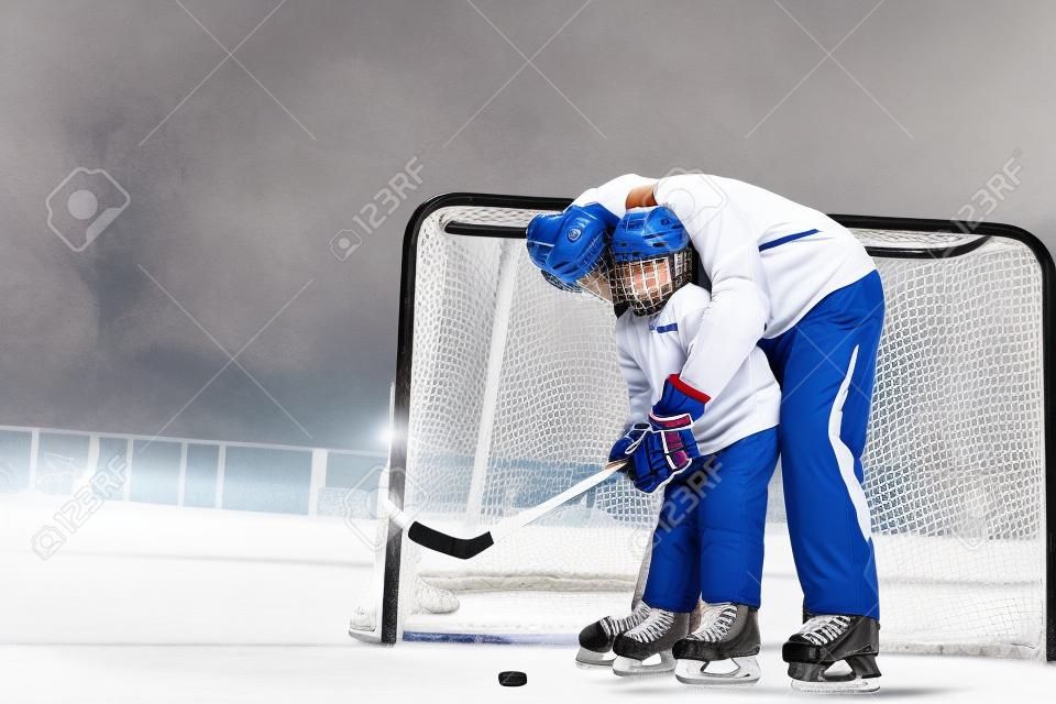 Father teach son to play ice hockey and hold hockey stick standing near gates on ice