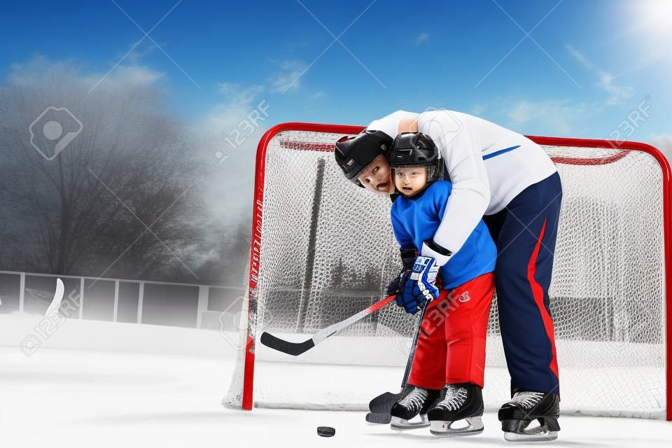 Father teach son to play ice hockey and hold hockey stick standing near gates on ice
