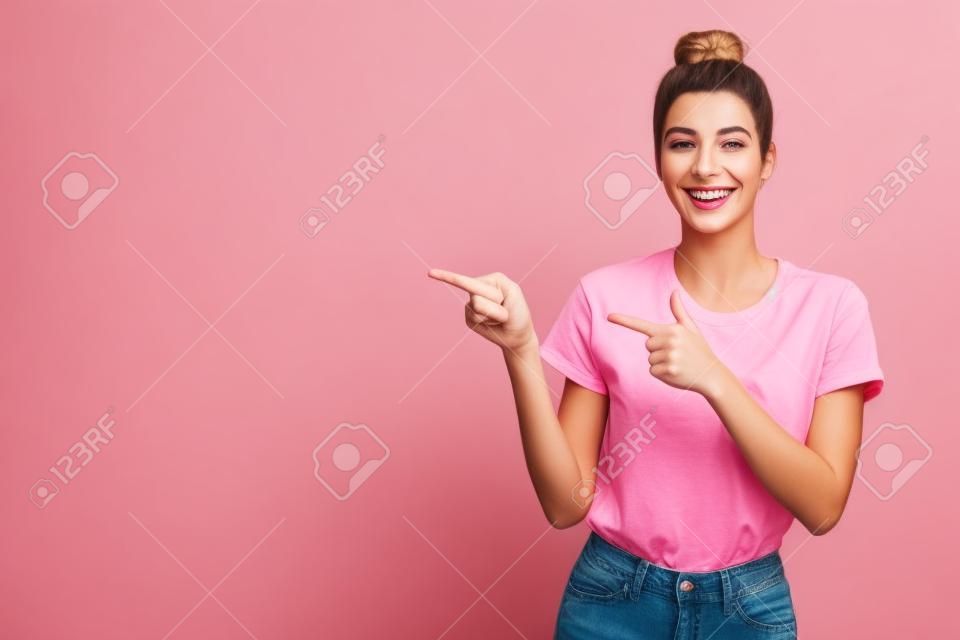 ピンクのTシャツとジーンズの見栄えの良い幸せな若いカリスマ的なヨーロッパの女性のウエストアップショットは、人差し指で左を指し、素晴らしいコピースペースを示す笑顔の印象を受けました