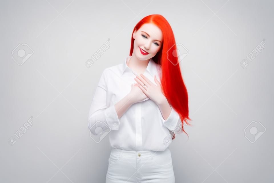 Manager di giovane donna con i capelli rossi e la pelle pallida, tenendo le mani sul cuore e ringraziandoti, essendo grato, esprimendo gratitudine in piedi su sfondo bianco