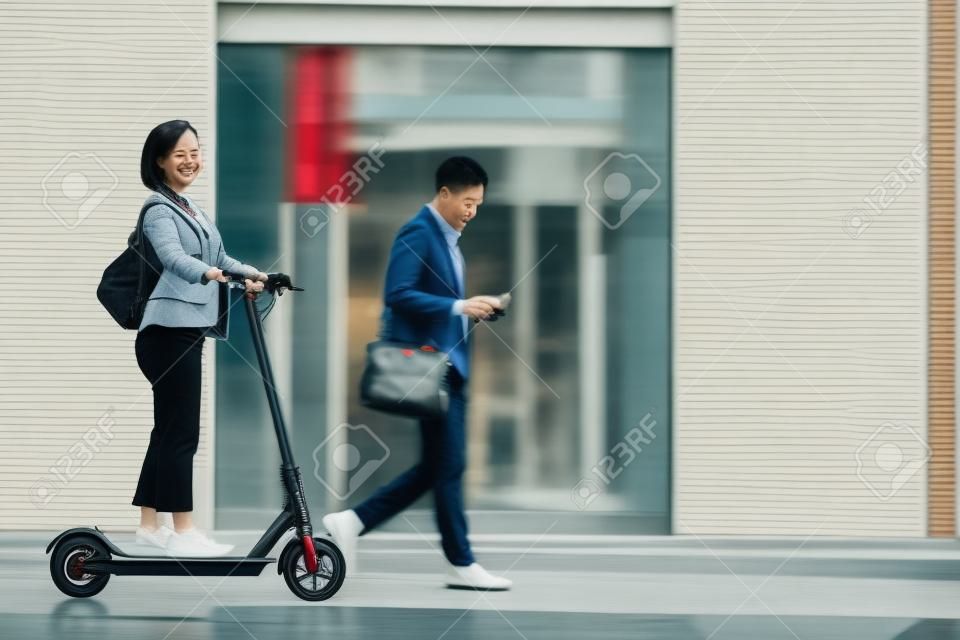都市で通勤する人々の完全な長さの肖像画は、前景に電動スクーターに乗ってアジアの女性に焦点を当て、スペースをコピー