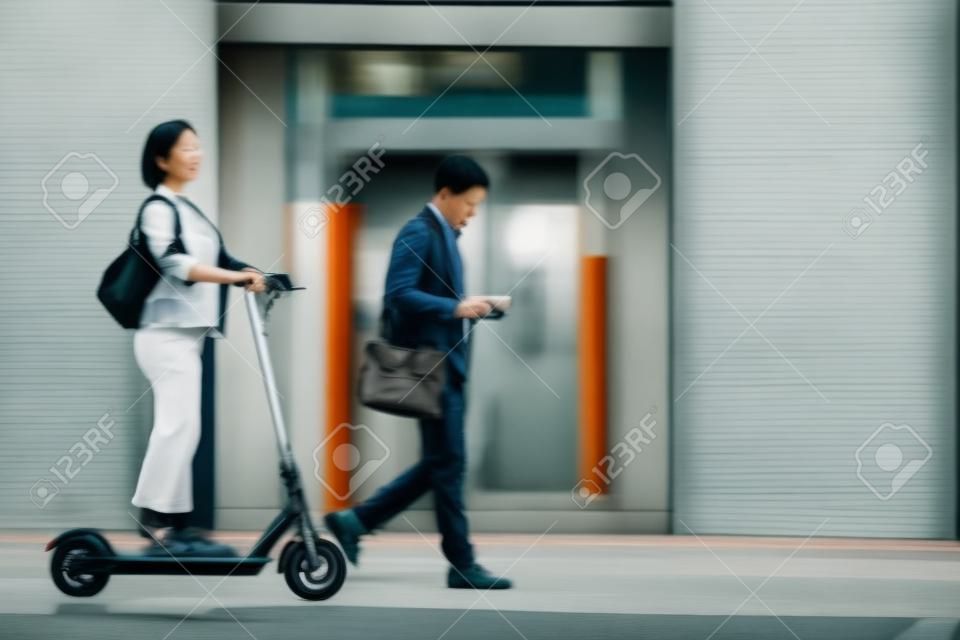 都市で通勤する人々の完全な長さの肖像画は、前景に電動スクーターに乗ってアジアの女性に焦点を当て、スペースをコピー