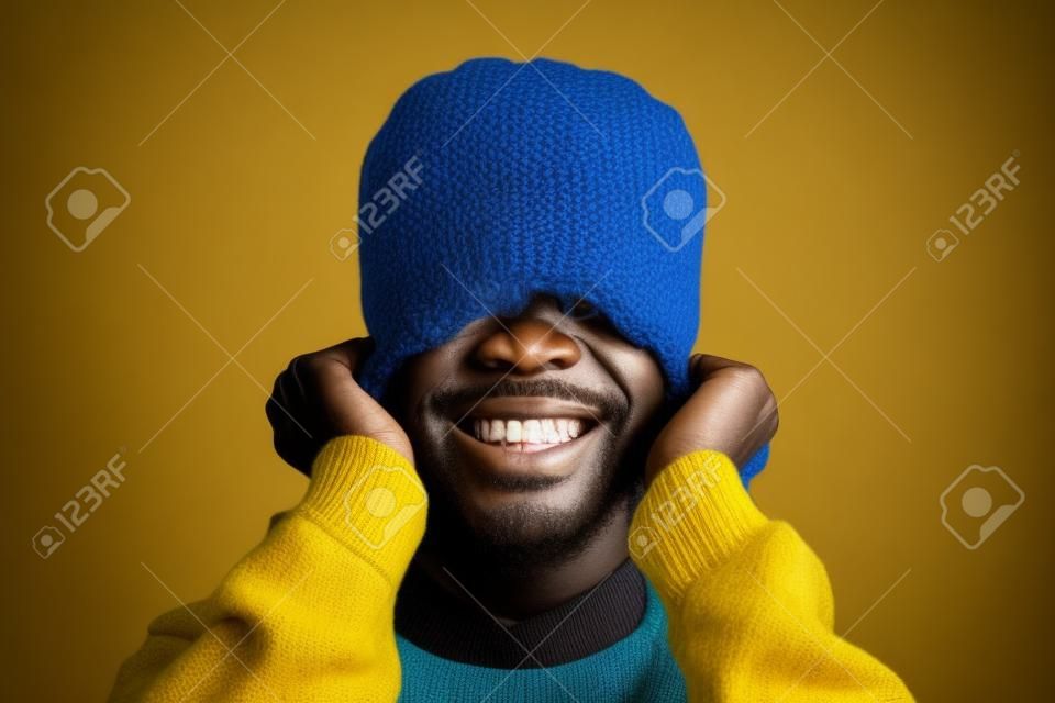 Retrato de un hombre africano emotivo sonriendo brillantemente a la cámara mientras tira de un sombrero de punto amarillo posando sobre fondo azul, copiando espacio
