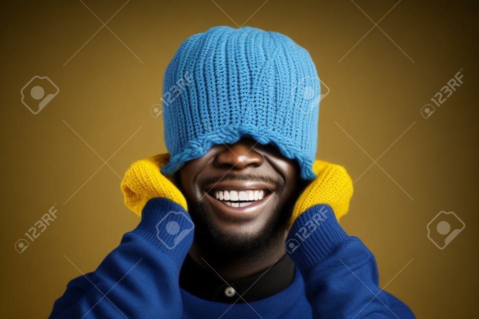 Retrato de un hombre africano emotivo sonriendo brillantemente a la cámara mientras tira de un sombrero de punto amarillo posando sobre fondo azul, copiando espacio