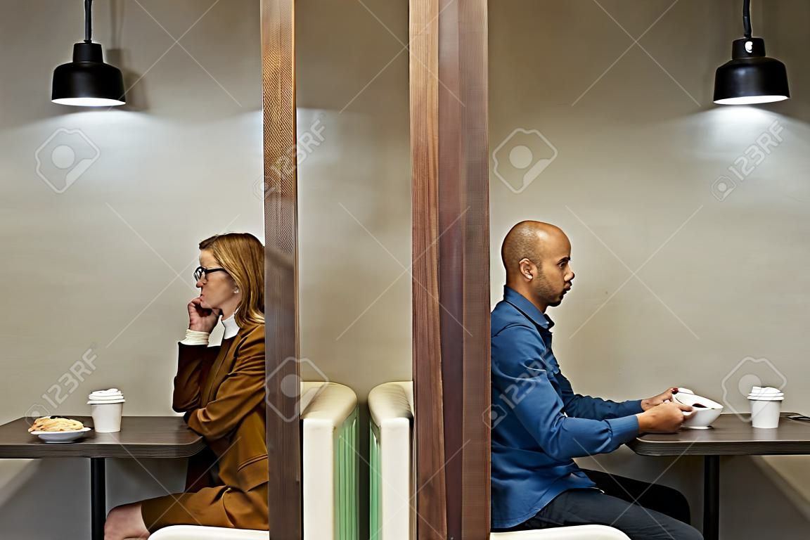 Minimales Seitenansichtsporträt von zwei erwachsenen Personen, die durch eine Wand getrennt sind, während sie in separaten Cafékabinen sitzen, Kopierraum